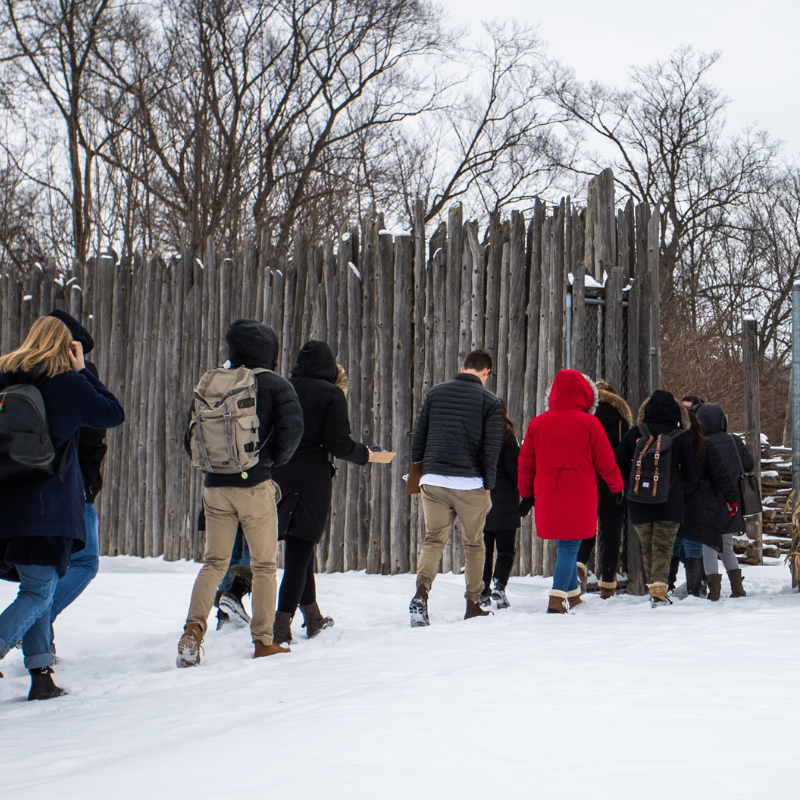 Several people walk in a line to enter a wooded area.