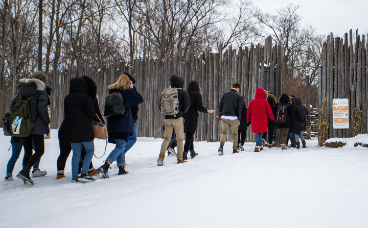 A group of people walk in a line to enter a wooded area.