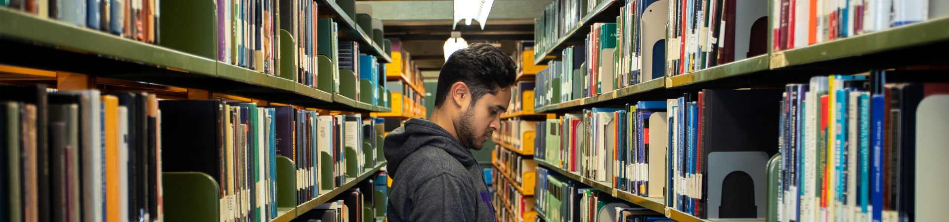 A young man reads a book in a library aisle.