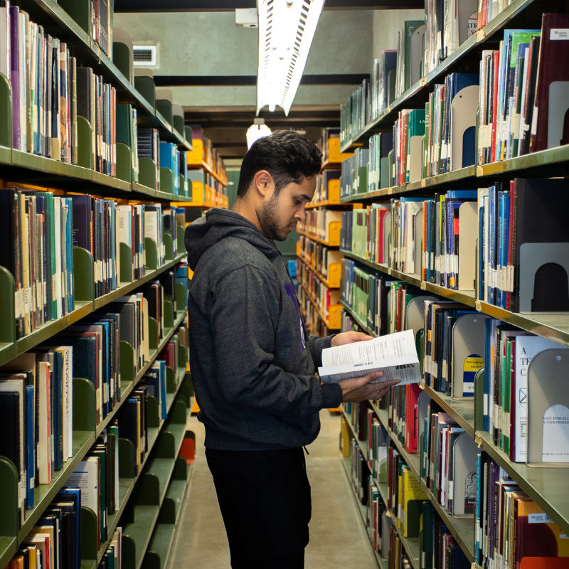 A young man reads a book in a library aisle.