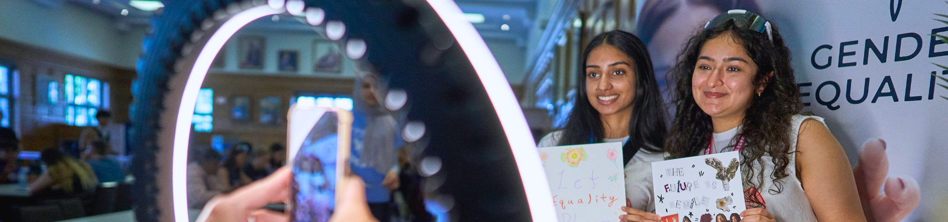 Two young women take a picture behind a ring light.